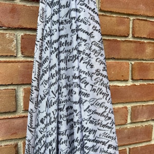May include: A white scarf with black lettering of various cities around the world. The lettering is in a handwritten style.