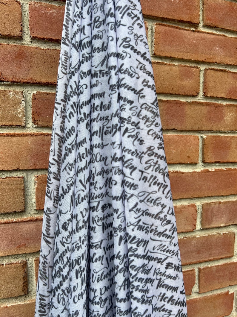 May include: A white scarf with black lettering of various cities around the world. The lettering is in a handwritten style.
