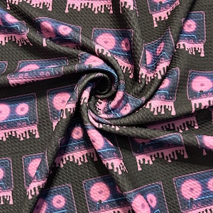 May include: Black fabric with a repeating pattern of pink cassette tapes with neon blue accents. The tapes are dripping pink and have a textured look.