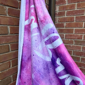 May include: A soft, pink and purple fleece blanket with a white "Dream" inscription. The blanket has a white, fluffy backing.