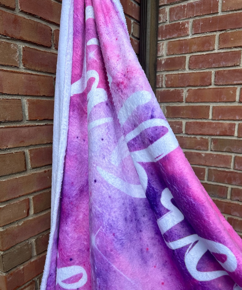 May include: A soft, pink and purple fleece blanket with a white "Dream" inscription. The blanket has a white, fluffy backing.