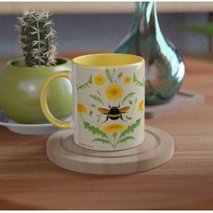 May include: A ceramic mug with a bright yellow interior and handle. The exterior features a repeating botanical illustration of yellow dandelion flowers, green leaves, and a central bumblebee with black and yellow stripes on a cream background.