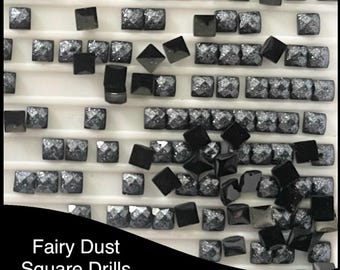 May include: A tray filled with square, black and gray Fairy Dust Square Drills, DMC 310. The drills are arranged in rows and columns, with some scattered around the tray. The drills have a faceted, sparkling appearance.