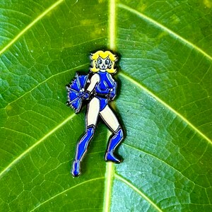 Puede incluir: Un pin de esmalte azul y amarillo con un personaje femenino de pelo rubio, que lleva un traje azul y sostiene un paraguas azul.