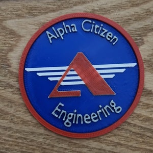 May include: A round patch with a blue center, red border, and white and red design. The text "Alpha Citizen Engineering" is arched around the top and bottom of the circle. The design includes a red triangle and white lines.