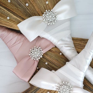 May include: Three wedding brooms with white, pink, and ivory satin bows and pearl-embellished brooches. The brooms are made of natural straw and have white pearl accents.