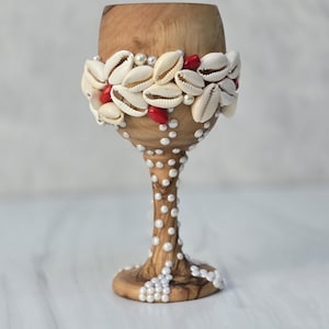 May include: A decorative wooden wine glass adorned with seashells, pearls, and red beads. The glass features a natural wood finish, with a band of seashells around the bowl and pearl accents on the stem and base. A unique, handcrafted item.