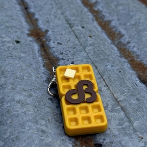 K&ouml;nnte beinhalten: Ein gelber, waffelf&ouml;rmiger Schl&uuml;sselanh&auml;nger mit einem braunen Sirup-Design und einem Butterst&uuml;ck. Der Schl&uuml;sselanh&auml;nger ist an einem silbernen Metallverschluss befestigt. Die Waffel hat ein Gittermuster.