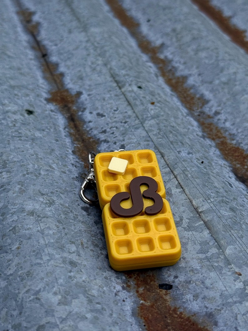 K&ouml;nnte beinhalten: Ein gelber, waffelf&ouml;rmiger Schl&uuml;sselanh&auml;nger mit einem braunen Sirup-Design und einem Butterst&uuml;ck. Der Schl&uuml;sselanh&auml;nger ist an einem silbernen Metallverschluss befestigt. Die Waffel hat ein Gittermuster.