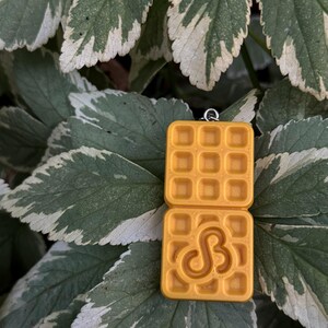 K&ouml;nnte beinhalten: Gelber Waffelanh&auml;nger mit quadratischer Form und Waffelmuster. Die untere Waffel hat ein stilisiertes "S"-Design. Der Anh&auml;nger h&auml;ngt an einer silberfarbenen Schlaufe. Der Hintergrund ist eine Mischung aus gr&uuml;nen und wei&szlig;en Bl&auml;ttern.
