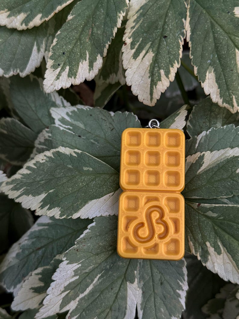 K&ouml;nnte beinhalten: Gelber Waffelanh&auml;nger mit quadratischer Form und Waffelmuster. Die untere Waffel hat ein stilisiertes "S"-Design. Der Anh&auml;nger h&auml;ngt an einer silberfarbenen Schlaufe. Der Hintergrund ist eine Mischung aus gr&uuml;nen und wei&szlig;en Bl&auml;ttern.