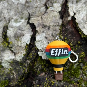 May include: A yellow hot air balloon keychain with a rainbow stripe design and the word "Effin" in white letters. The keychain has a silver ring and a brown basket. The background is a textured tree trunk.