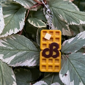 K&ouml;nnte beinhalten: Ein gelber Waffel-Schl&uuml;sselanh&auml;nger mit einem Butterst&uuml;ck und einem schokoladenfarbenen "dB"-Design. Der Schl&uuml;sselanh&auml;nger ist an einer silberfarbenen Kette und einem Verschluss befestigt. Der Hintergrund zeigt gr&uuml;ne und wei&szlig;e Bl&auml;tter.