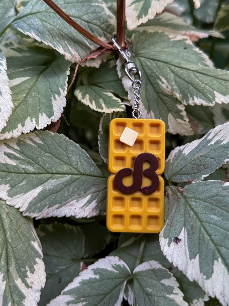 K&ouml;nnte beinhalten: Ein gelber Waffel-Schl&uuml;sselanh&auml;nger mit einem Butterst&uuml;ck und einem schokoladenfarbenen "dB"-Design. Der Schl&uuml;sselanh&auml;nger ist an einer silberfarbenen Kette und einem Verschluss befestigt. Der Hintergrund zeigt gr&uuml;ne und wei&szlig;e Bl&auml;tter.