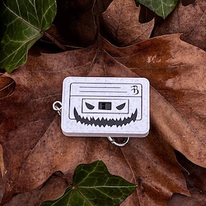 May include: White cassette tape keychain with a black and white grinning face design. The keychain is on a bed of brown and green leaves.