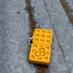K&ouml;nnte beinhalten: Ein gelber, waffelf&ouml;rmiger Schl&uuml;sselanh&auml;nger mit einem silbernen Verschluss. Ein Waffelquadrat hat ein stilisiertes "SB"-Design. Der Schl&uuml;sselanh&auml;nger besteht aus einem einfarbigen gelben Material und liegt auf einer verwitterten, grauen Oberfl&auml;che.