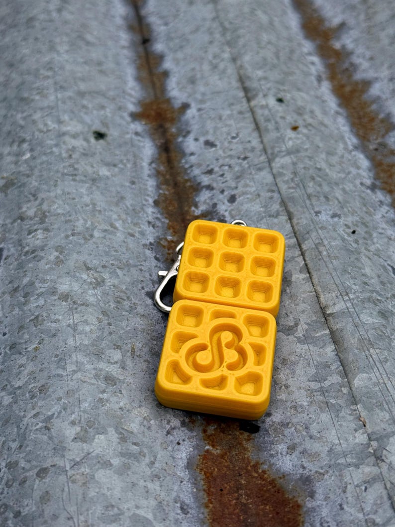 K&ouml;nnte beinhalten: Ein gelber, waffelf&ouml;rmiger Schl&uuml;sselanh&auml;nger mit einem silbernen Verschluss. Ein Waffelquadrat hat ein stilisiertes "SB"-Design. Der Schl&uuml;sselanh&auml;nger besteht aus einem einfarbigen gelben Material und liegt auf einer verwitterten, grauen Oberfl&auml;che.