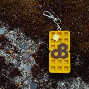 K&ouml;nnte beinhalten: Ein gelber Waffel-Schl&uuml;sselanh&auml;nger mit einem silbernen Verschluss. Die Waffel hat ein Butterst&uuml;ck und einen braunen Buchstaben "B". Der Schl&uuml;sselanh&auml;nger liegt auf einer moosigen Oberfl&auml;che.