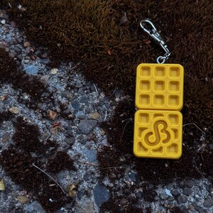 K&ouml;nnte beinhalten: Ein gelber, waffelf&ouml;rmiger Schl&uuml;sselanh&auml;nger mit einem silbernen Verschluss. Der Schl&uuml;sselanh&auml;nger hat oben und unten ein Waffelmuster mit einem stilisierten "SB"-Design auf der unteren H&auml;lfte. Der Hintergrund ist eine Mischung aus Moos und Stein.