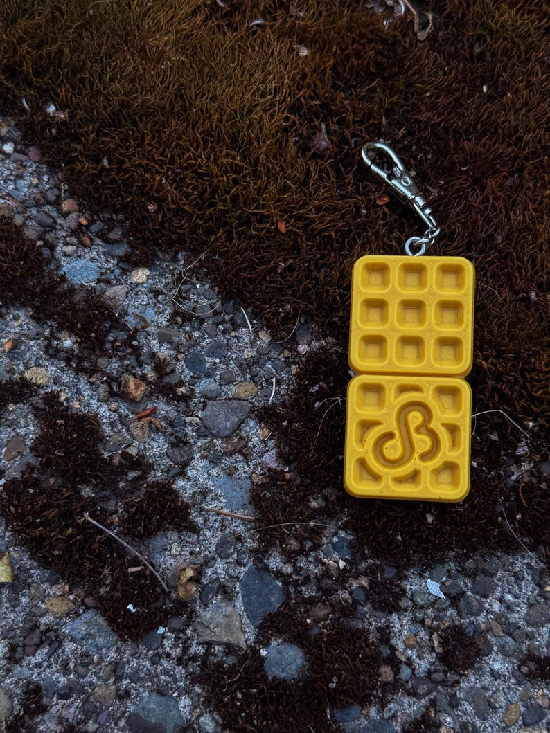 K&ouml;nnte beinhalten: Ein gelber, waffelf&ouml;rmiger Schl&uuml;sselanh&auml;nger mit einem silbernen Verschluss. Der Schl&uuml;sselanh&auml;nger hat oben und unten ein Waffelmuster mit einem stilisierten "SB"-Design auf der unteren H&auml;lfte. Der Hintergrund ist eine Mischung aus Moos und Stein.