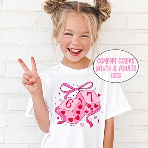 May include: White t-shirt featuring a pink graphic of two dice with the numbers 6 and 7, decorated with hearts and ribbons. The text "COMFORT COLORS YOUTH & ADULTS SIZES" is in a circle. The model is wearing black pants.