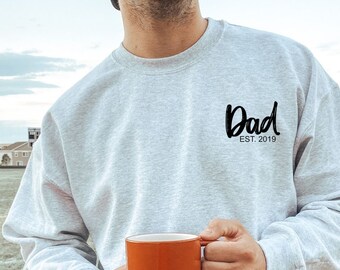 May include: A gray sweatshirt with the text "Dad Est. 2019" printed on the front. The person is holding a red mug.