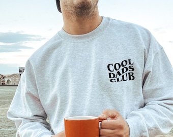 May include: A gray sweatshirt with the text "COOL DADS CLUB" in black lettering. The person wearing the sweatshirt is holding a red mug.