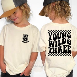 May include: Two children wearing cream-colored t-shirts and baseball caps. The front of one shirt has a black peace sign with the name "William" below it. The back of the other shirt says "Young Wild & Three" in black, with a checkered border.