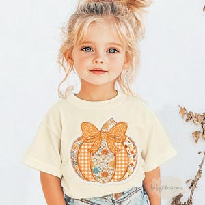 May include: Cream-colored t-shirt featuring a pumpkin design with a bow. The pumpkin has a gingham pattern, floral accents, and a lace trim. The shirt is worn by a young child.