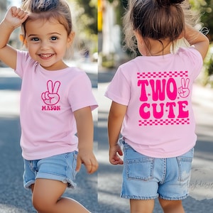 May include: A pink t-shirt with a peace sign and the name "MASON" in pink. The back of the shirt says "TWO CUTE" in pink. The child is wearing denim shorts and white shoes.