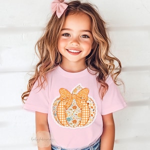 May include: A light pink t-shirt featuring a pumpkin design with a bow. The pumpkin is decorated with floral and plaid patterns in orange, yellow, and white. The shirt is worn by a young child with long, wavy brown hair and a pink bow.