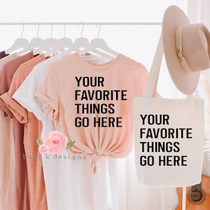May include: A peach-colored t-shirt and a white tote bag both feature the text "Your Favorite Things Go Here". The t-shirt is tied at the front and the tote bag is hanging from a wooden rack. A light brown hat is hanging from the tote bag.