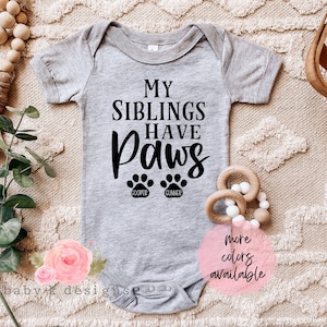 May include: Gray baby onesie with the text "My siblings have paws" and two paw prints with the names "Cooper" and "Cummer" underneath. The onesie is on a beige background with a wooden toy and a pink flower.
