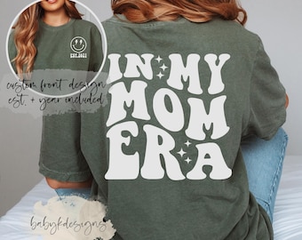 May include: A green t-shirt with the text "IN MY MOM ERA" in white, retro-style lettering with stars. The text is on the back of the shirt.