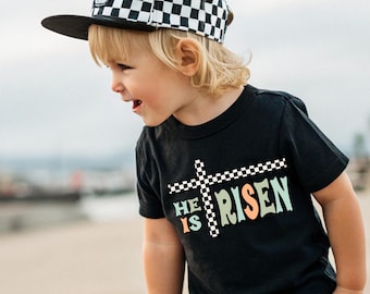May include: A young child wearing a black t-shirt with the text "He is Risen" in colorful letters and a checkered black and white baseball cap. The text is displayed over a checkered black and white cross.