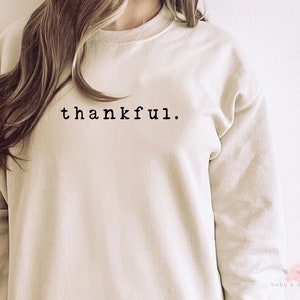 Op de afbeelding: Cr&egrave;mekleurige sweatshirt met het woord "thankful" in zwart gedrukt, gecentreerd bovenaan. De sweatshirt heeft een ronde hals en lange mouwen. De afbeelding is een close-up van het bovenlichaam.