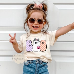 May include: A young child wearing a light beige t-shirt with a ghost graphic that says "BOO" in checkered purple and orange letters. The ghost is wearing a pink bow and holding a small orange pumpkin.