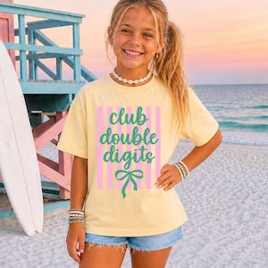 May include: A pale yellow short-sleeve t-shirt featuring a graphic print of vertical pink stripes with the green text club double digits and a green bow illustration centered on the front. The shirt has a crew neckline and a relaxed fit.
