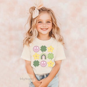 May include: A young girl wearing a white t-shirt with a green, yellow, and pink design of shamrocks, flowers, and smiley faces. The shirt has the text "babydesigns" on the bottom.