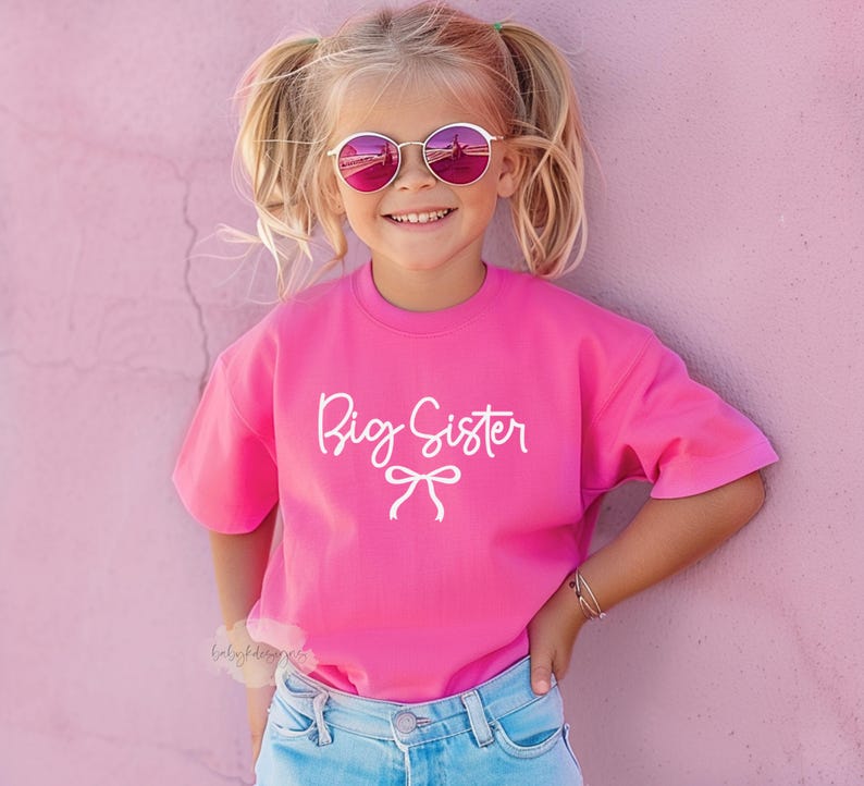 May include: A young girl wearing a bright pink t-shirt with the words "Big Sister" in white script and a bow graphic. She is also wearing pink sunglasses and blue jeans. The background is a solid pink wall.