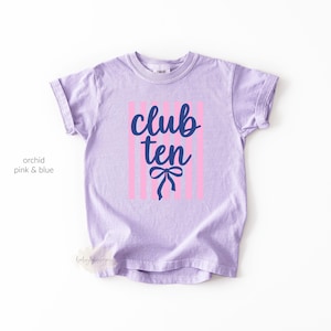 May include: An orchid-colored short-sleeve t-shirt featuring a graphic design with vertical light pink stripes. Centered on the stripes is the dark blue text "club ten" in a cursive script font, positioned above a dark blue graphic bow icon.