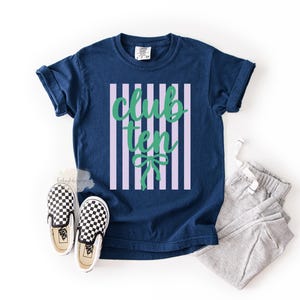 May include: A navy blue short-sleeve t-shirt featuring a central graphic of vertical light purple stripes with the green text "club ten" and a green bow icon. The shirt is paired with a pair of gray sweatpants and black and white checkered slip-on shoes.
