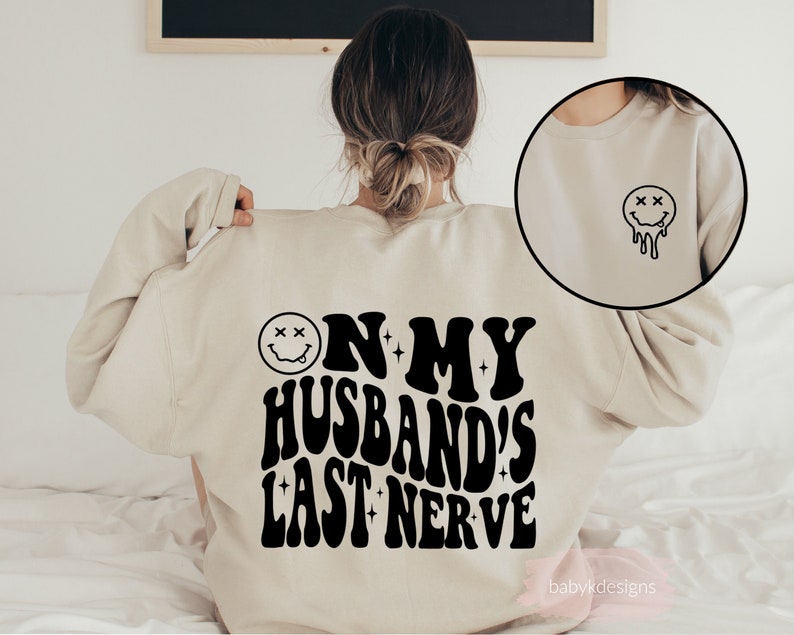 May include: A beige sweatshirt with the text "On My Husband's Last Nerve" in black, with a dripping smiley face graphic. The sweatshirt is being worn by a person.