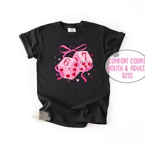May include: Black t-shirt featuring a graphic of two pink dice with hearts and the numbers 6 and 7. A pink ribbon and the text "Comfort Colors Youth & Adults Sizes" are also visible.
