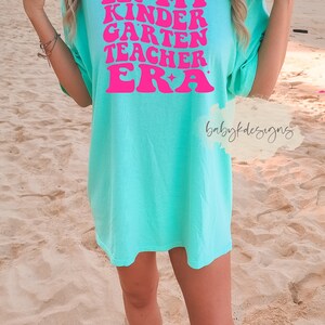 May include: A turquoise t-shirt with the text "IN MY KINDERGARTEN TEACHER ERA" in pink retro font.