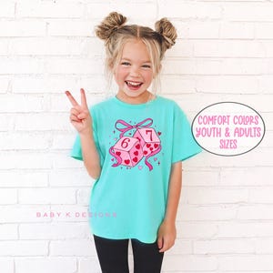 May include: A turquoise t-shirt featuring a graphic of two pink dice with hearts and the numbers 6 and 7. The dice are tied with a pink bow. The text "Comfort Colors Youth & Adults Sizes" is in a circle.