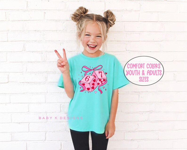 May include: A turquoise t-shirt featuring a graphic of two pink dice with hearts and the numbers 6 and 7. The dice are tied with a pink bow. The text "Comfort Colors Youth & Adults Sizes" is in a circle.