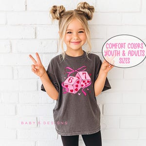 May include: A dark gray t-shirt featuring a graphic of two pink dice with hearts and the numbers 6 and 7. The dice are tied with a pink bow and ribbons. The text "Comfort Colors Youth & Adults Sizes" is in a pink oval.