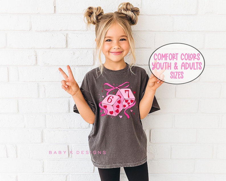 May include: A dark gray t-shirt featuring a graphic of two pink dice with hearts and the numbers 6 and 7. The dice are tied with a pink bow and ribbons. The text "Comfort Colors Youth & Adults Sizes" is in a pink oval.