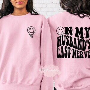 May include: Pink sweatshirt with a dripping smiley face on the front and the text "On my husband's last nerve" on the back.